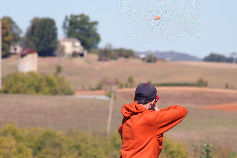 A shooter in an orange hoodie tracking a clay target